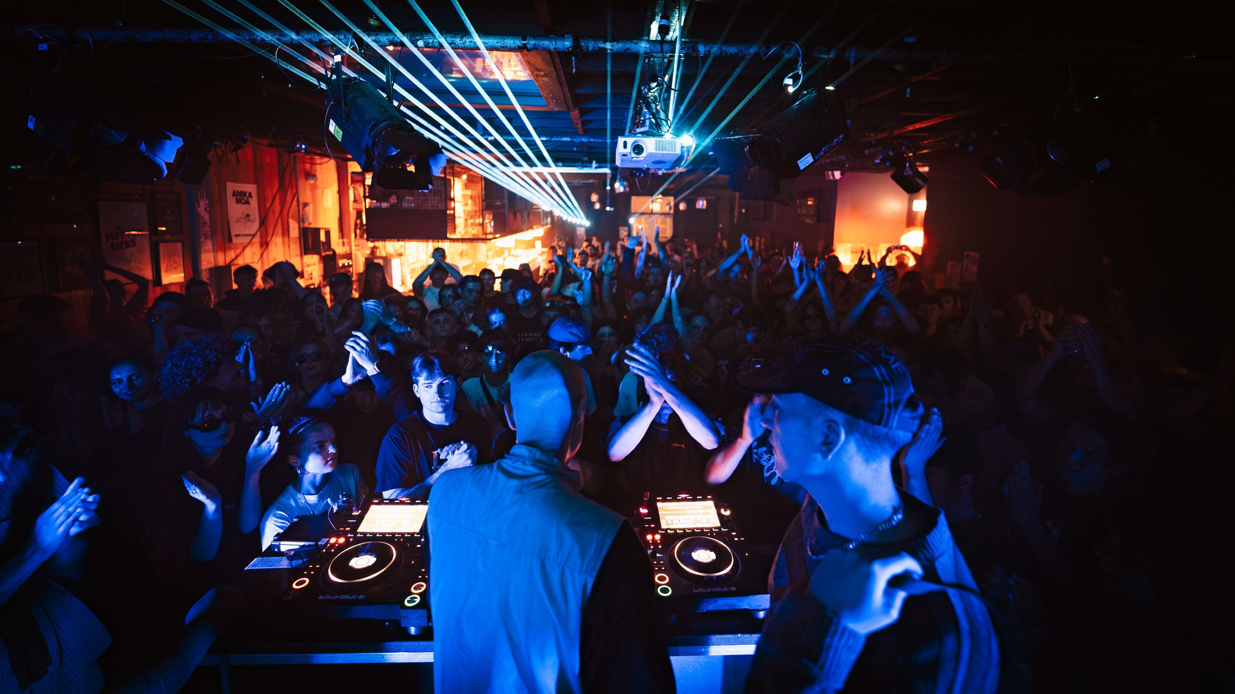 A shot taken from behind the DJ, looking over their head at a pumping crowd. Lasers cross the air above them.